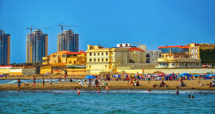 Belebter Stadtstrand mit Sonnenbädern und Schwimmern vor einer modernen Hochhäuserskyline an einem strahlenden Sommertag