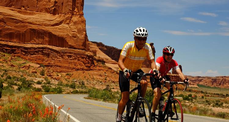 Close-up of two cyclists pedalling past dramatic orange rock walls under a bright blue sky.