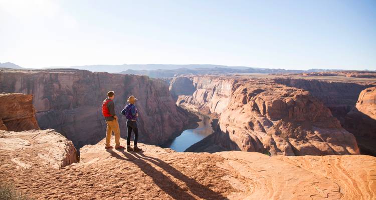 Two hikers stand on the rim overlooking the dramatic bend of the Colorado River far below.