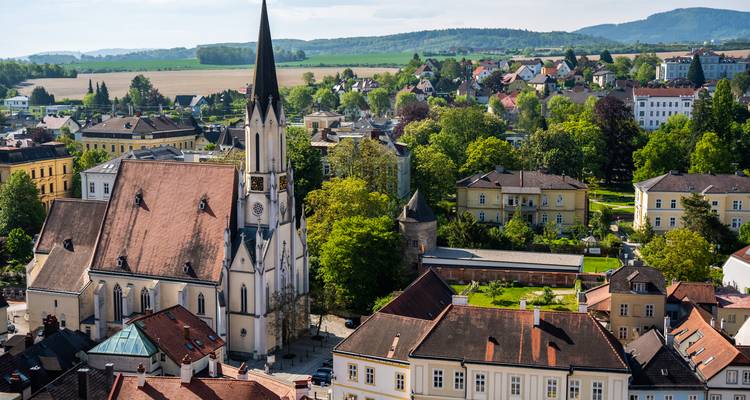 Charmante österreichische Stadt mit hohem Kirchturm, eingebettet in grüne Felder und bewaldete Hügel.