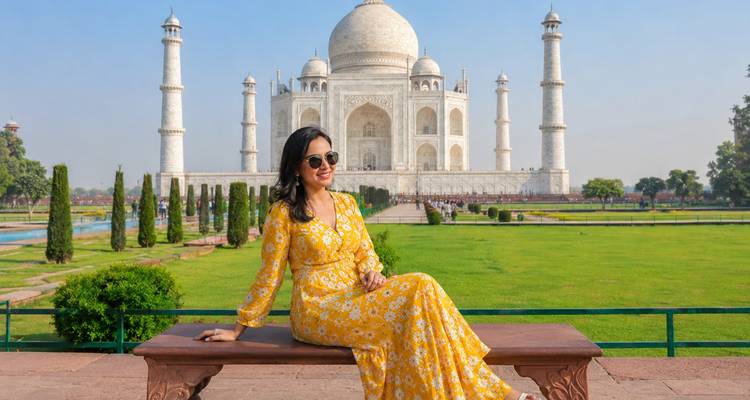 Mujer joven con un vestido amarillo brillante sentada en un banco con una vista clara del Taj Mahal detrás de ella.