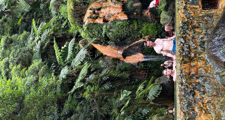 Les baigneurs se détendent dans une petite piscine naturelle d'eau chaude située sous une cascade thermale aux traînées orangées, au cœur d'une jungle dense.