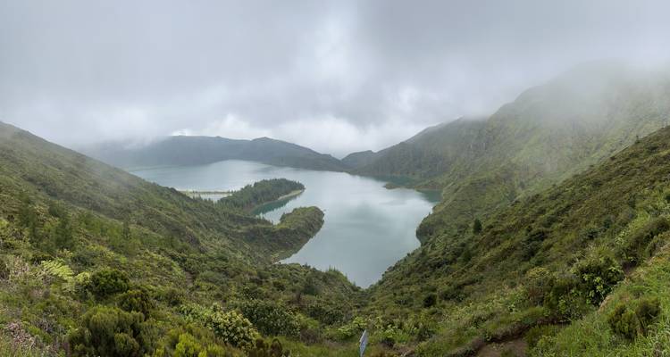 Vue panoramique large d'un lac de cratère brumeux entouré de pentes raides et verdoyantes.