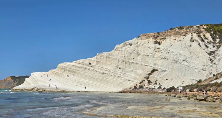 Visitors explore base of striking white stepped cliff beside shallow coastal waters.