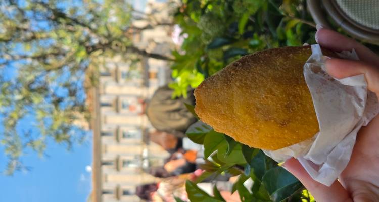Close-up of hand holding a golden fried arancino wrapped in paper with blurred cityscape behind.