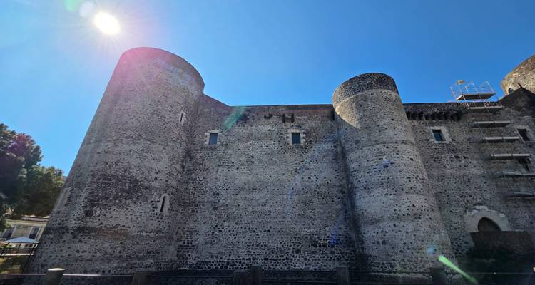 Massive stone fortress with cylindrical towers lit by midday sun and lens flare against a deep blue sky.