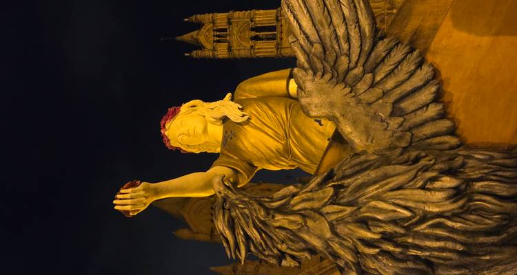 Illuminated night-time statue of a crowned female figure with outstretched arm beside a large winged eagle.