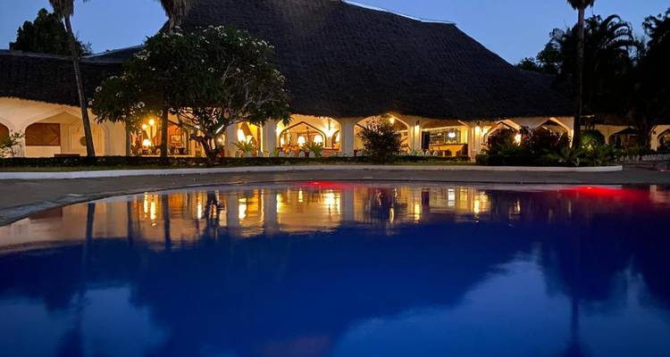 Evening view of thatched safari lodge lit warmly beside a calm outdoor pool reflecting lights.