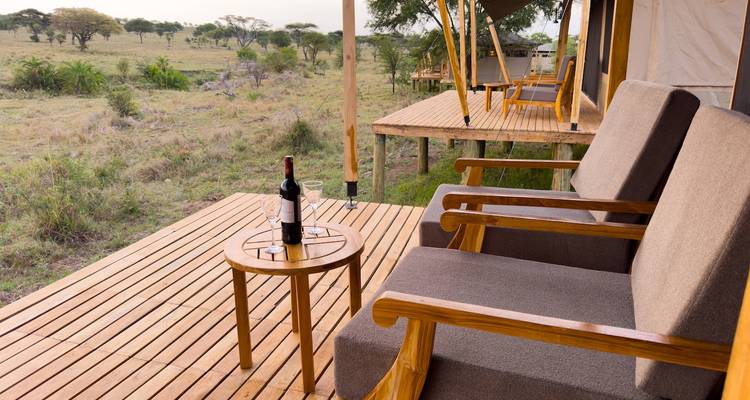 Terraza de madera con sillas cómodas y una botella de vino con vistas a la sabana abierta desde un campamento de tiendas.