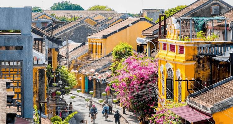 Flower-lined street of yellow heritage buildings in Hoi An ancient town bustling with pedestrians.