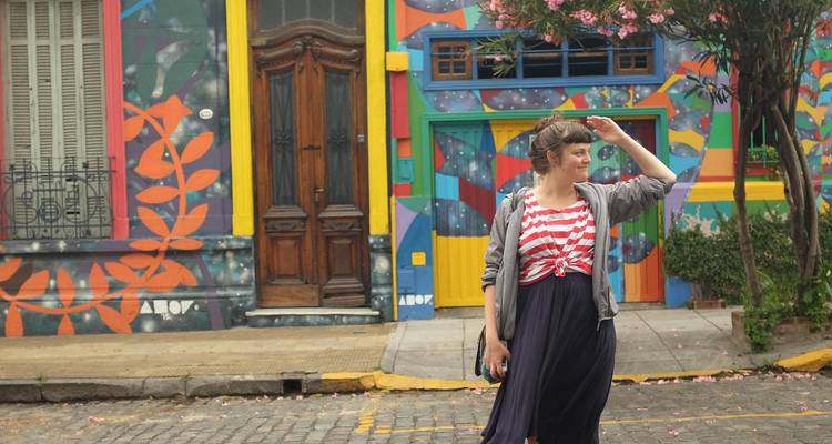 Una mujer joven con ropa casual sonríe y se protege los ojos mientras está de pie en una calle empedrada llena de murales pintados con colores vibrantes y puertas.