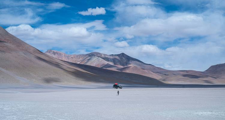Vaste saline d'altitude avec un véhicule solitaire et un randonneur écrasés par les pics lointains.