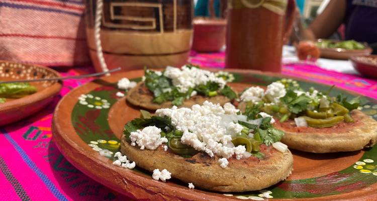 Nahaufnahme von traditionellen mexikanischen Sopes, belegt mit frischem Koriander, Käse und Gemüse auf einem bemalten Tonteller über einem leuchtenden Stoff.