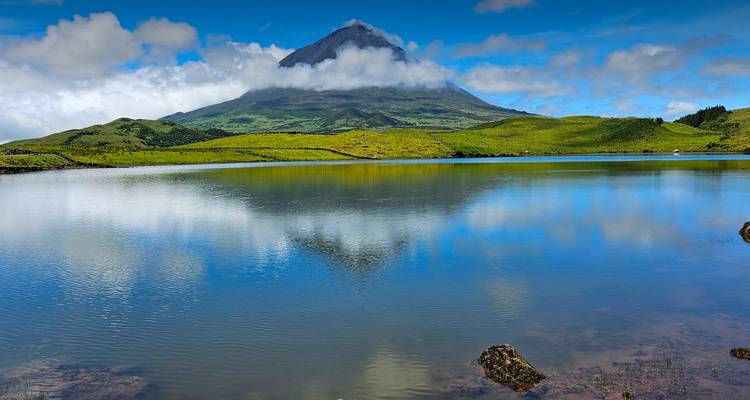 Le volcan Pico aux pics enneigés parfaitement reflété dans un lac calme sous un ciel bleu éclatant et des nuages épars.