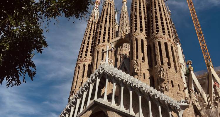 Angle ascendant de la façade de la Nativité de la Sagrada Familia encadré par des feuilles d'arbres et un ciel bleu.