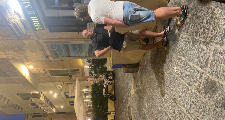 Deux hommes discutent sur une ruelle étroite pavée de pierre, bordée de bâtiments historiques et illuminée par les lumières des terrasses en soirée.