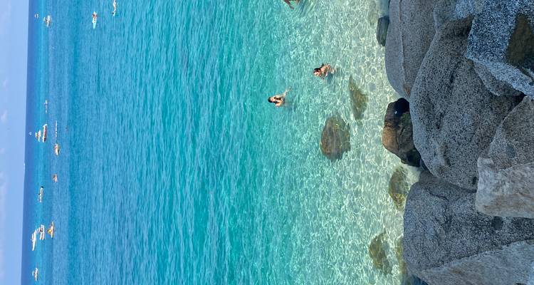 Eau turquoise cristalline où les nageurs profitent de la mer près d'une côte rocheuse et de bateaux amarrés.