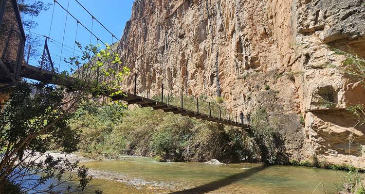 Passerelle suspendue étroite surplombant une rivière peu profonde avec des parois rocheuses imposantes.
