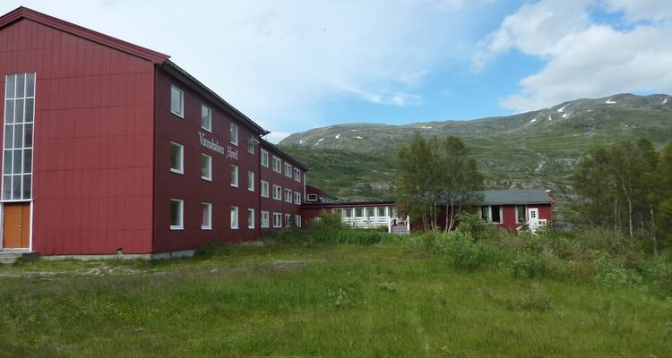Chalet alpin rouge situé sur une prairie herbacée sous un terrain montagneux vallonné et un ciel partiellement nuageux.