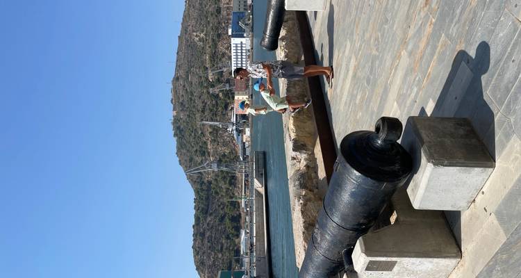 Promenade portuaire avec canons historiques où un parent soulève un enfant près de l'eau