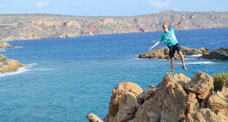 Young man leaping from jagged coastal rocks with a vivid blue sea and cliffs behind.