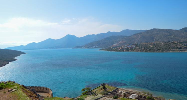 Wide turquoise bay bordered by mountains and scattered villages viewed from above stone ruins.