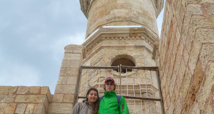 Two travellers standing at the base of a historic stone lighthouse tower against a cloudy sky.