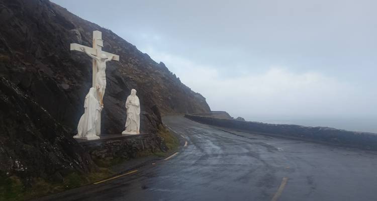 Statue de crucifixion au bord de la route surplombant une route côtière escarpée enveloppée de brume.