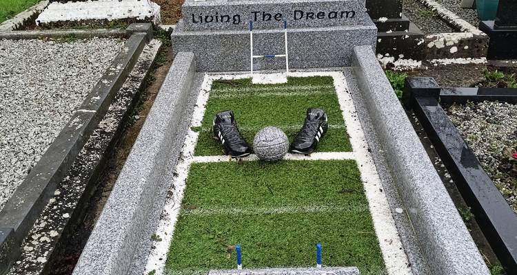 Una tumba única con estilo de mini cancha de deportes con botas y pelota, lápida que dice 'Living The Dream'.