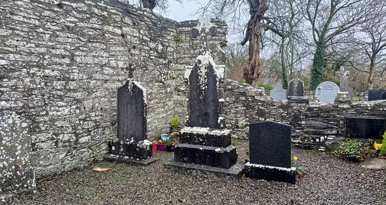 Muro de piedra desgastada que rodea lápidas oscuras manchadas de líquenes en un tranquilo cementerio rural.