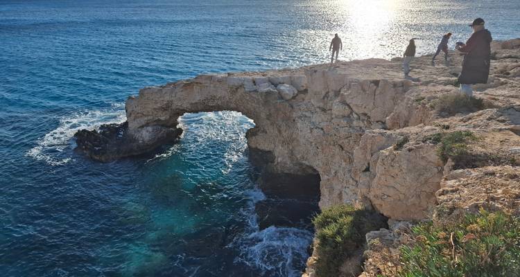 People explore a natural sea bridge at sunset with glittering water below