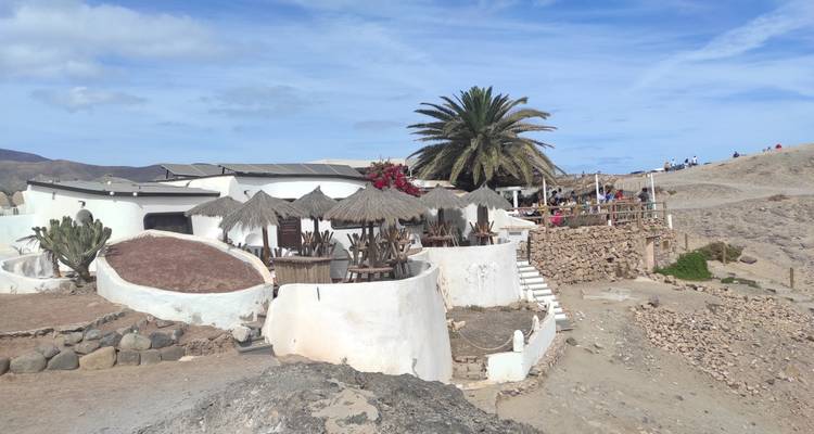 Bar de plage aux murs blanchis à la chaux avec des parasols en chaume, un palmier et des visiteurs se détendant sous un ciel bleu.