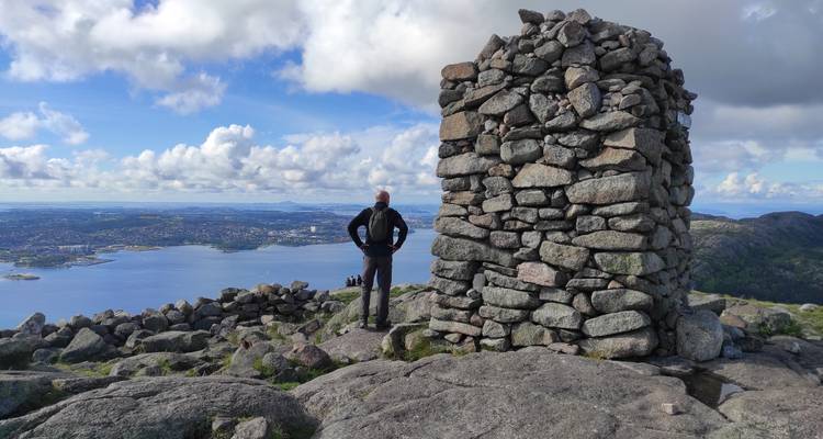 Randonneur debout à côté d'un haut cairn en pierre sur un sommet montagneux surplombant une ville côtière et un fjord.