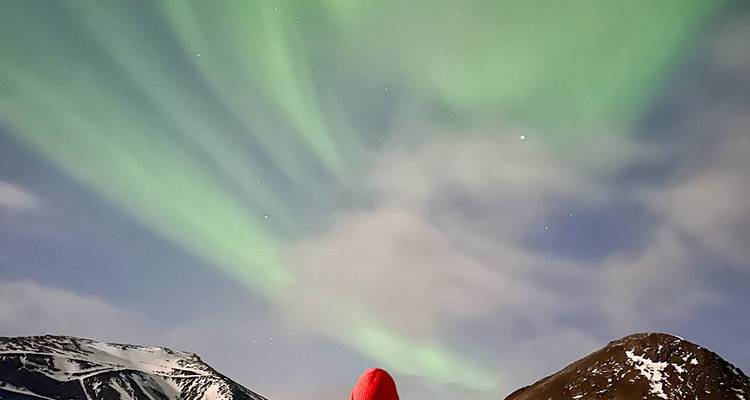 Une personne portant un chapeau rouge regarde vers le haut les aurores boréales d'un vert éclatant au-dessus de montagnes enneigées.