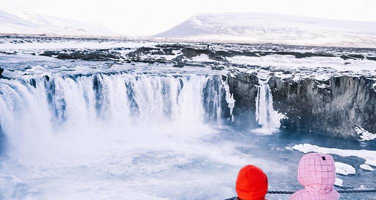 Deux enfants portant des chapeaux colorés admirent la puissante cascade gelée de la cascade de Goðafoss.