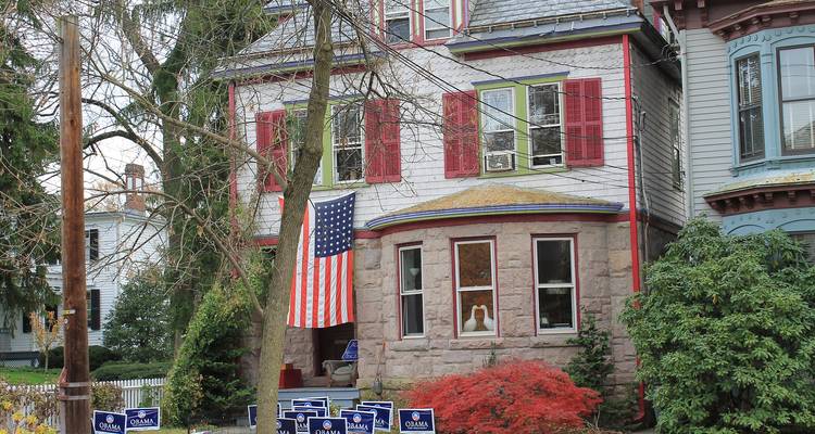 Maison à façade en pierre avec volets rouges, grand drapeau américain et pancartes politiques de jardin parmi les arbres d'automne.