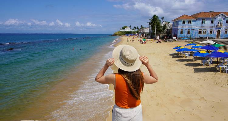 Vue de dos d'une femme tenant un chapeau de soleil tout en se tenant debout sur une large plage de sable fin avec une eau turquoise et des parasols.