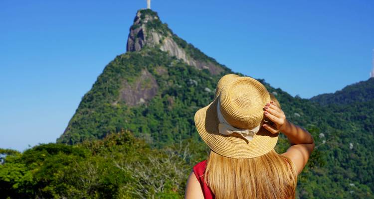 Femme portant un chapeau de paille regardant vers la statue emblématique du Christ Rédempteur au sommet d'un pic verdoyant et luxuriant sous un ciel bleu dégagé.
