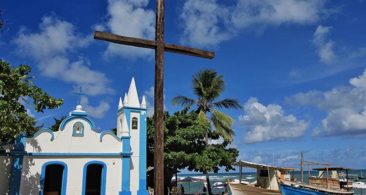 Petite chapelle en bord de mer avec des bordures bleues, une grande croix en bois, un palmier et des bateaux de pêche sur un ciel bleu éclatant.