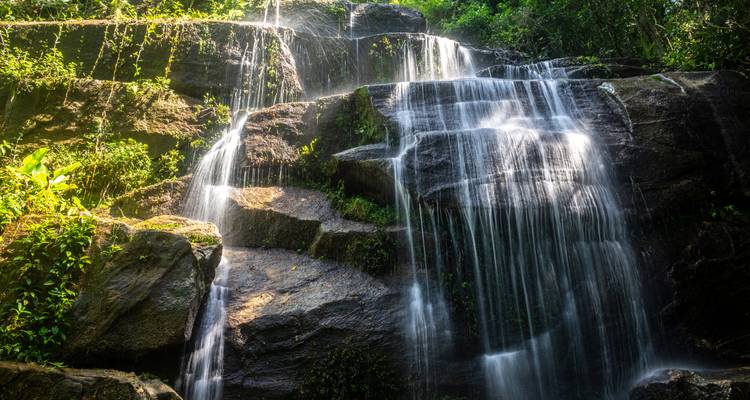 Cascade de forêt tropicale à plusieurs niveaux s'écoulant sur des rochers sombres avec un effet d'eau fluide et soyeuse.