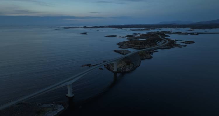 Vue crépusculaire aérienne d'un pont sinueux reliant de petits îlots rocheux à travers une mer calme et sombre.