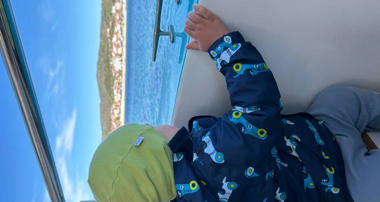A child in a colorful jacket gazes out from a boat railing toward a coastal town and blue water.