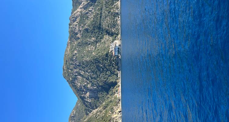 A remote seaside monastery perches at the base of rugged mountains rising from vivid blue sea.