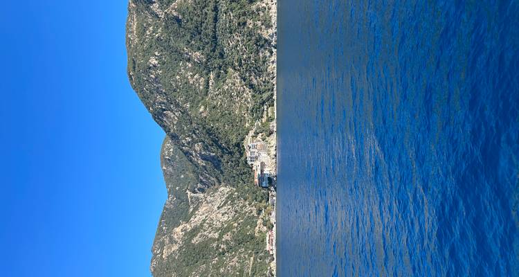 A historic monastery complex clings to steep green mountains above deep blue sea under cloudless skies.