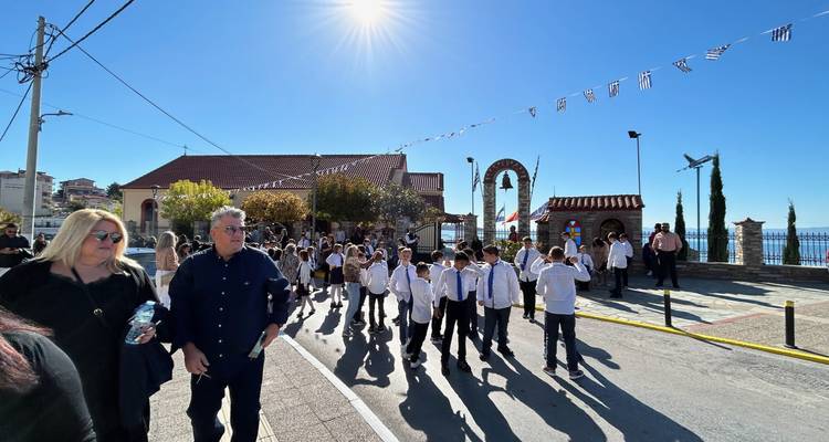 Children in uniform march in a sunny street parade while adults watch, with festive bunting overhead and sea glimpses beyond.