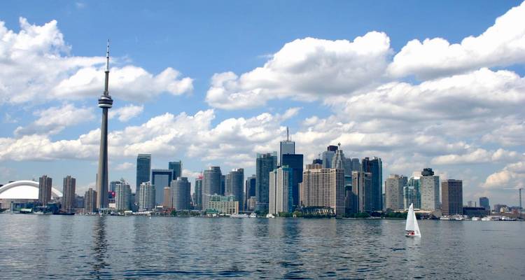 Panorama de la skyline de Toronto avec la Tour CN et un voilier blanc sur les eaux calmes du lac Ontario sous des nuages légers et cotonneux.
