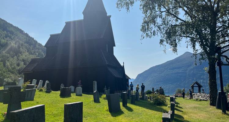 Église en bois à colombages baignée de soleil avec un petit cimetière devant, entourée de montagnes vertes et de ciel bleu.