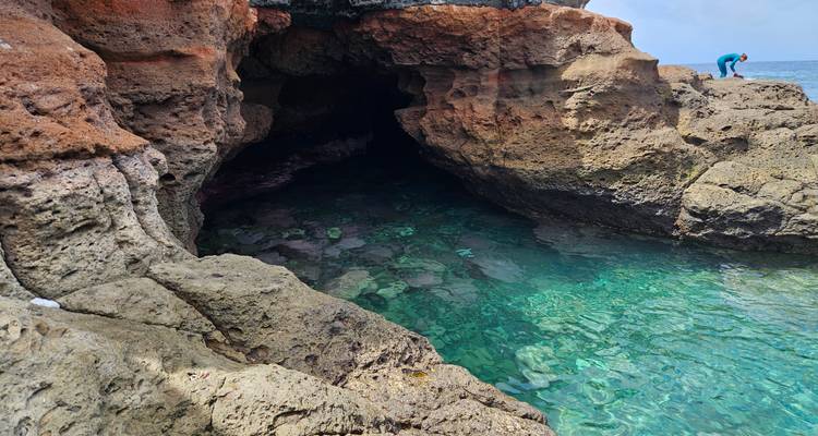 Eau turquoise cristalline à l'intérieur d'une grotte marine rocheuse tandis qu'une personne regarde vers le bas depuis une corniche au-dessus.