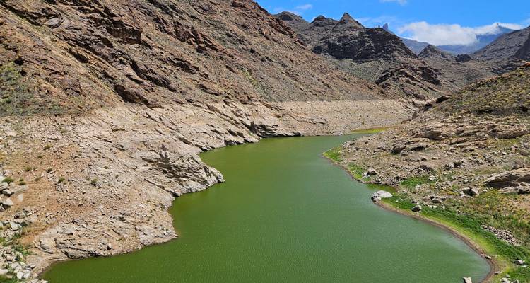 Réservoir étroit et verdoyant sinuant entre des collines rocheuses et arides sous un ciel bleu éclatant.