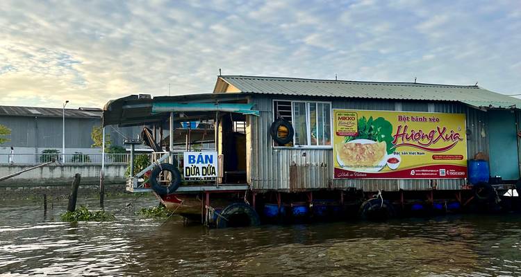 Floating food stall anchored on a river at dawn with a pastel sky in the background.